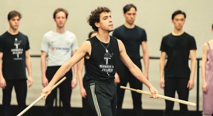 Dancer strides confidently across the stage with a weapon in each hand, is arms slightly spread threateningly