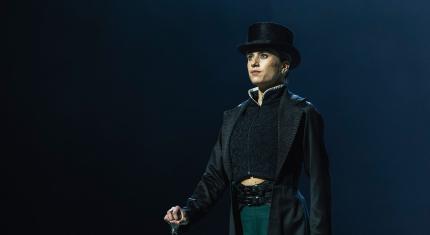 Standing proud, cane in one hand, hearing a tall hat and long coat, Anne Lister looks imperious