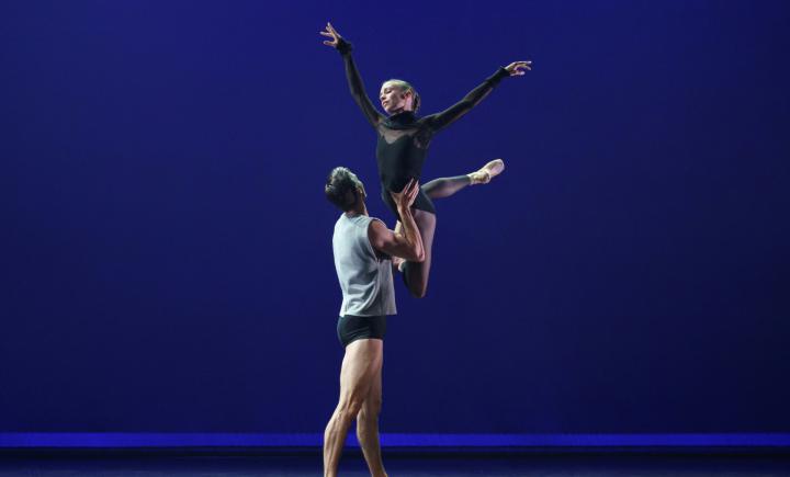 A female dancer in black leotard is lifted in the air by a male dance in black trunks and grey vest.