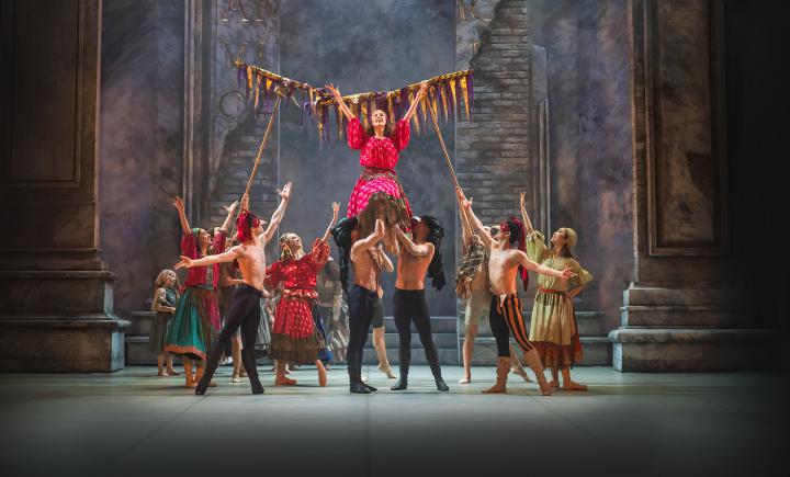 Men in masks and black leggings lift a woman in a pink patterned dress and she raises her arms in celebration