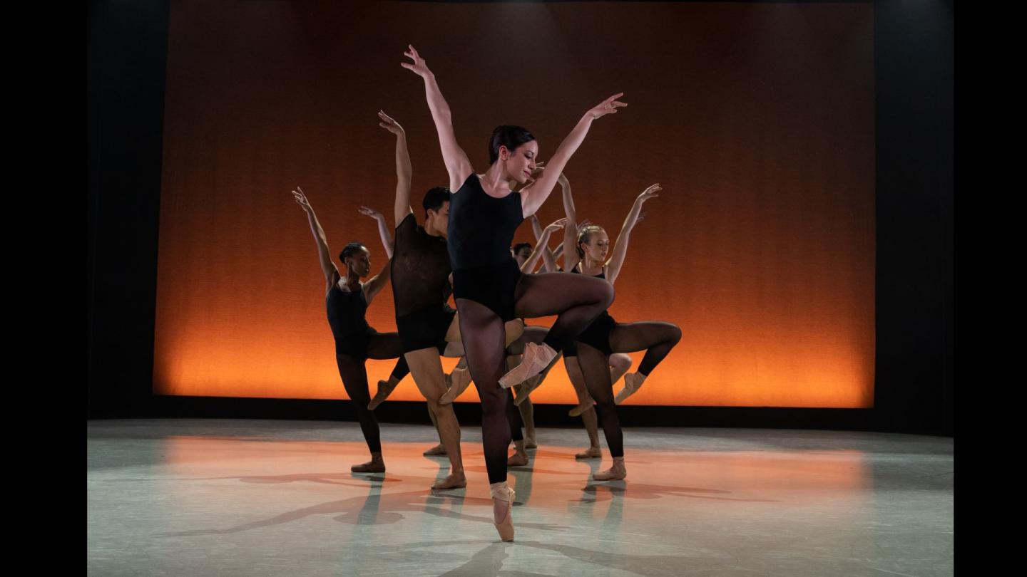 7 dancers stand en pointe, both arms in the air with a warm orange background behind them