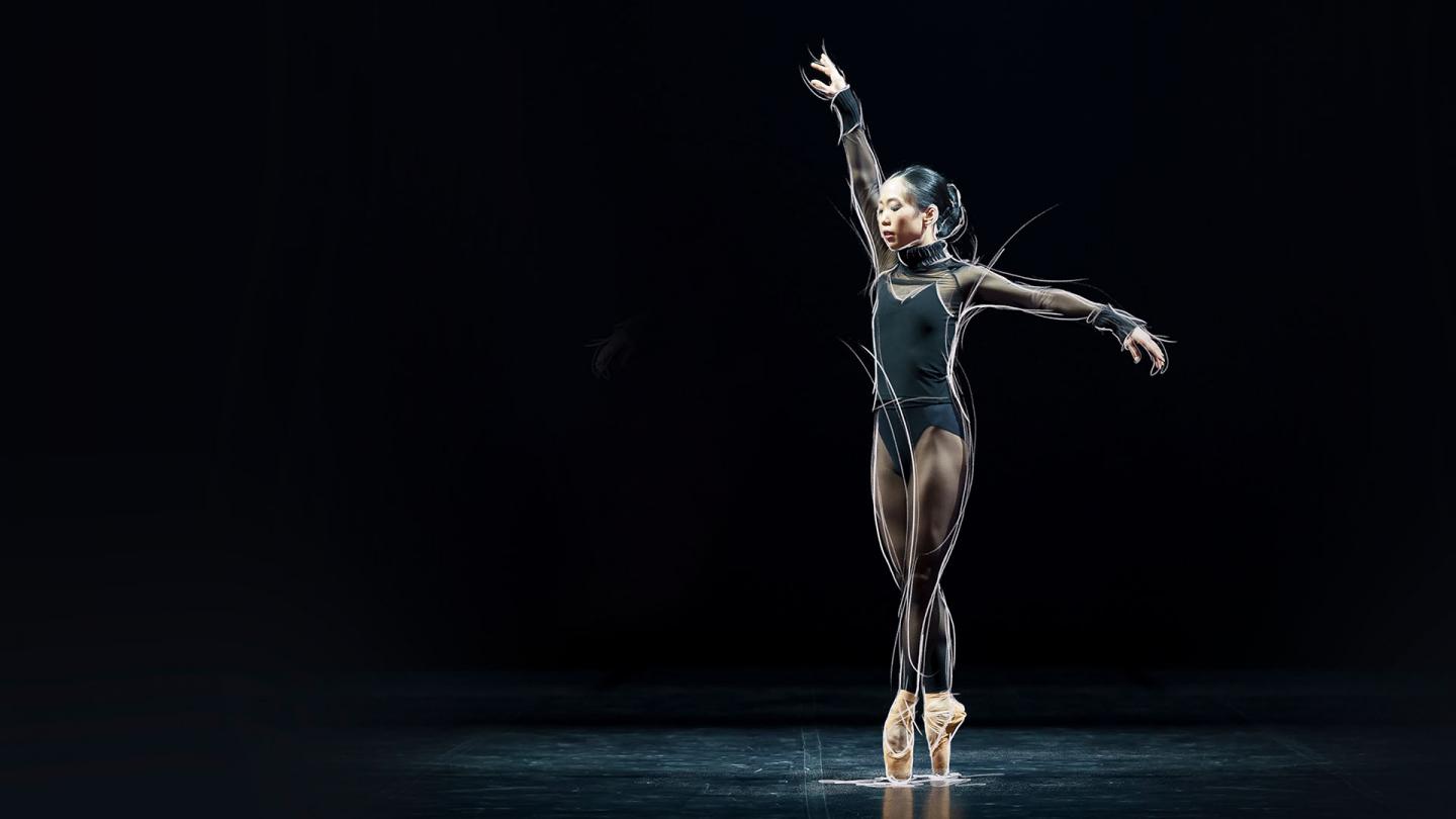 Dancer in a black leotard stands en pointe against a black background with one arm extended above her and one to her side.