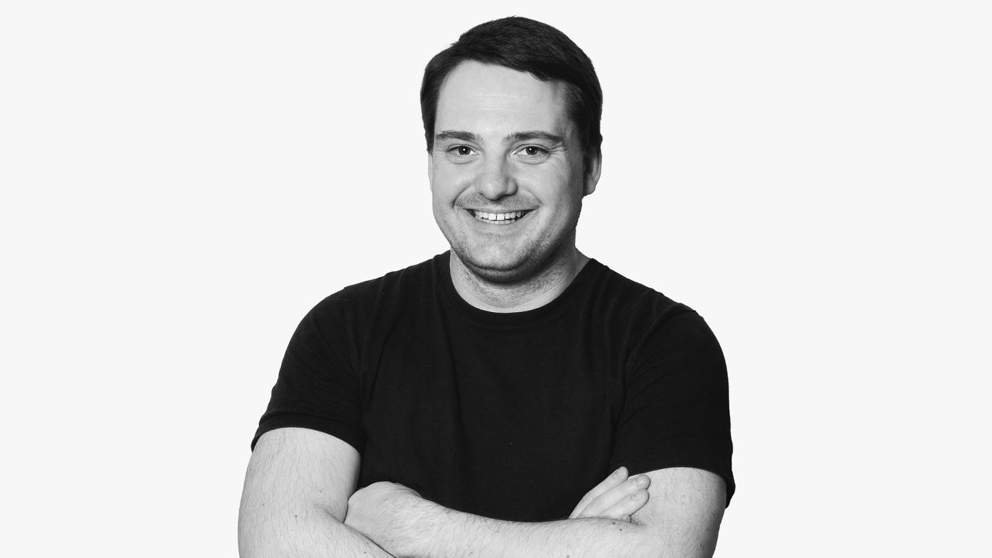 Black and white photograph of white man smiling, wearing a black T-shirt, arms folded across his chest.