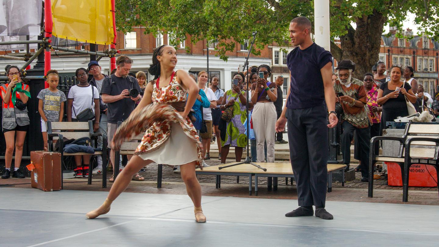 Two black dancers, one in a bright flower dress, the other in dark trousers and T-shirt, dance on stage while members of the public look on