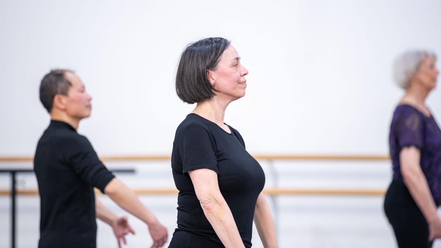 Woman in profile, wearing black T-shirt with darker bobbed hair, striking classic ballet pose in class