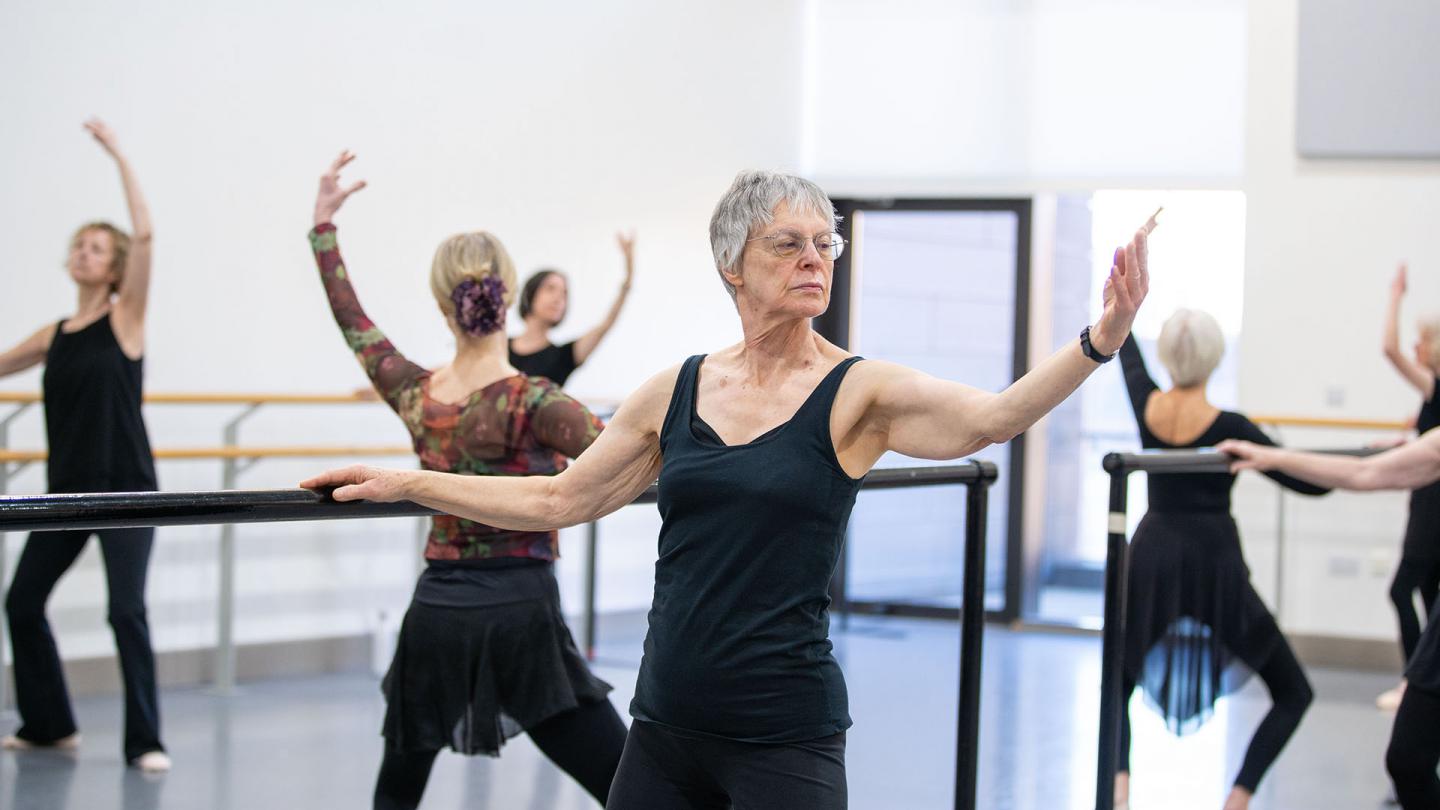 Class of dancers at the barre, one hand holding on for balance, the other arm held out, palm up forearm raised, knees slightly bent