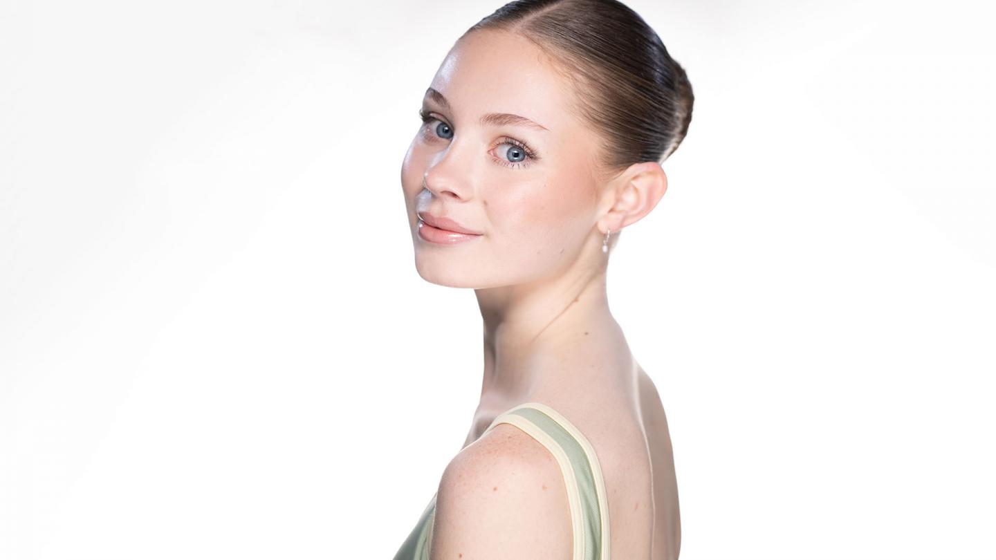 Dancer smiling, looking over shoulder directly at the camera, light tight back hair, green leotard top
