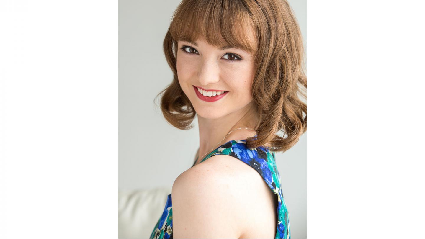 Woman with long auburn hair, wearing a blue patterned top looks over her shoulder at the camera, smiling