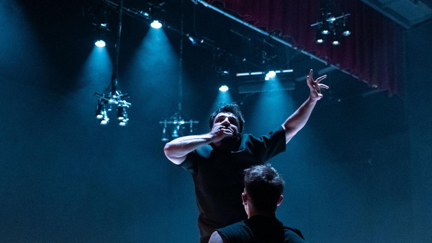 Two dancers stand on a dark stage lit by intense blue spotlights. One dancer stands with his back to the camera, while the other faces forward, covering his mouth with one hand and raising one arm in an expressive gesture.