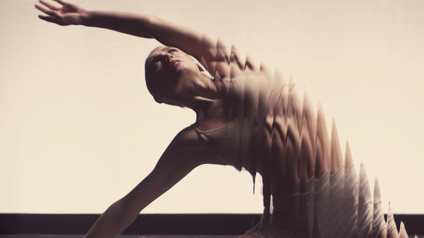 Woman in a dance floor with a cream backdrop, holding herself steadily on the ground with one arm and has the other above her head while looking up