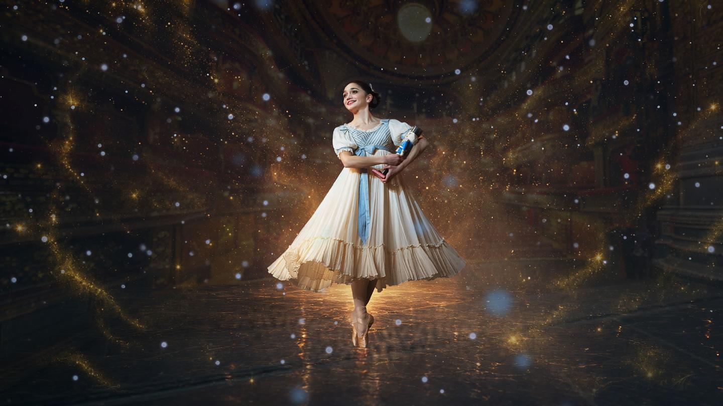 A dancer in Edwardian costume hold a Nutcracker doll in her arms, stands en pointe and smiles with childlike wonder