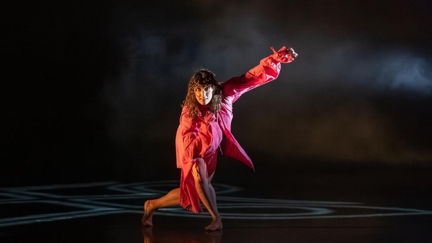 On a dark stage, a woman in pink is pictured. She has her right leg planted on the floor with her left leg crossing behind it, making her lunge. She is looking forwards with her left arm up in the air and her right close to her chest.