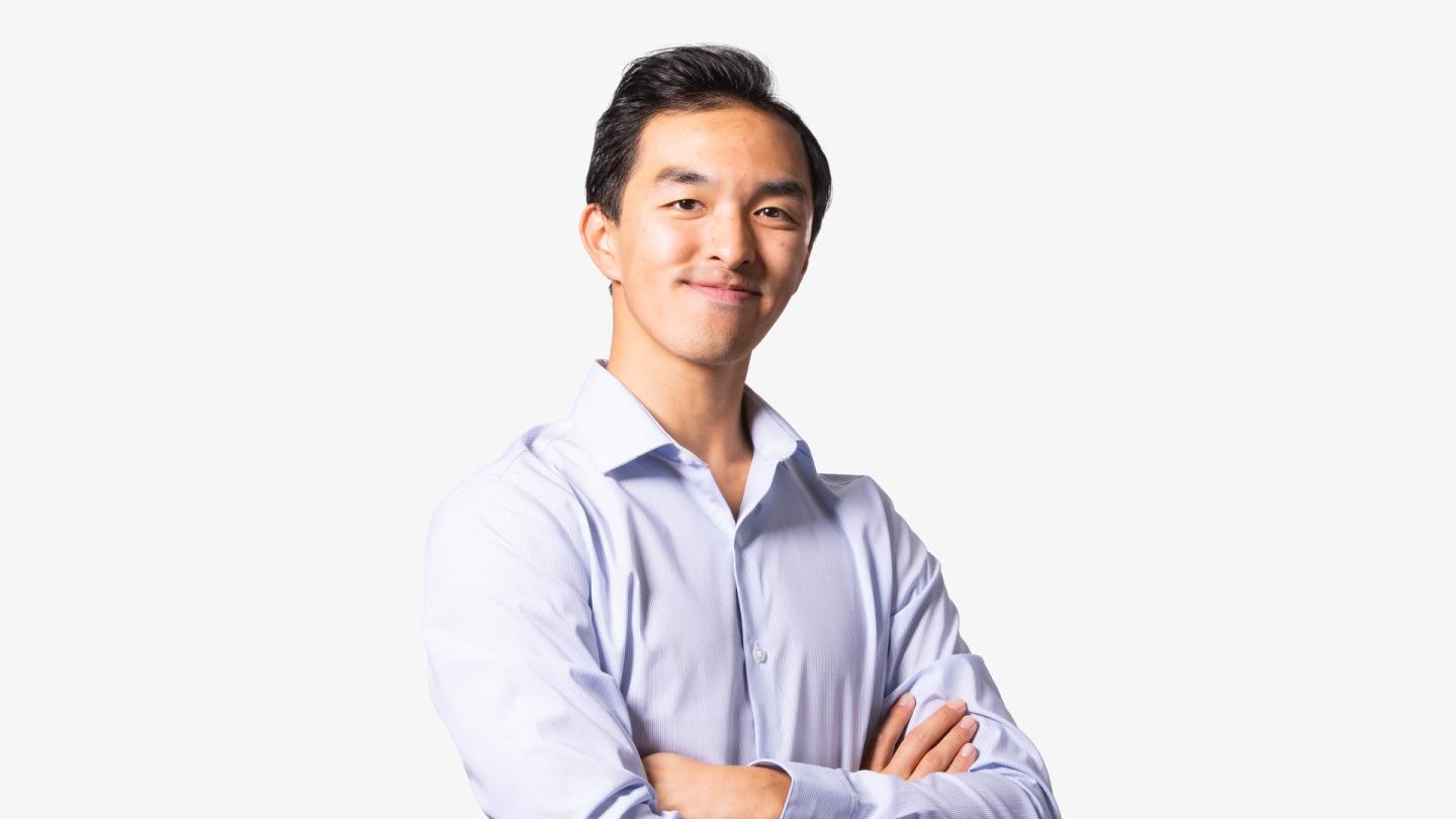 A man with dark hair stood in with arms crossed in a light purple shirt