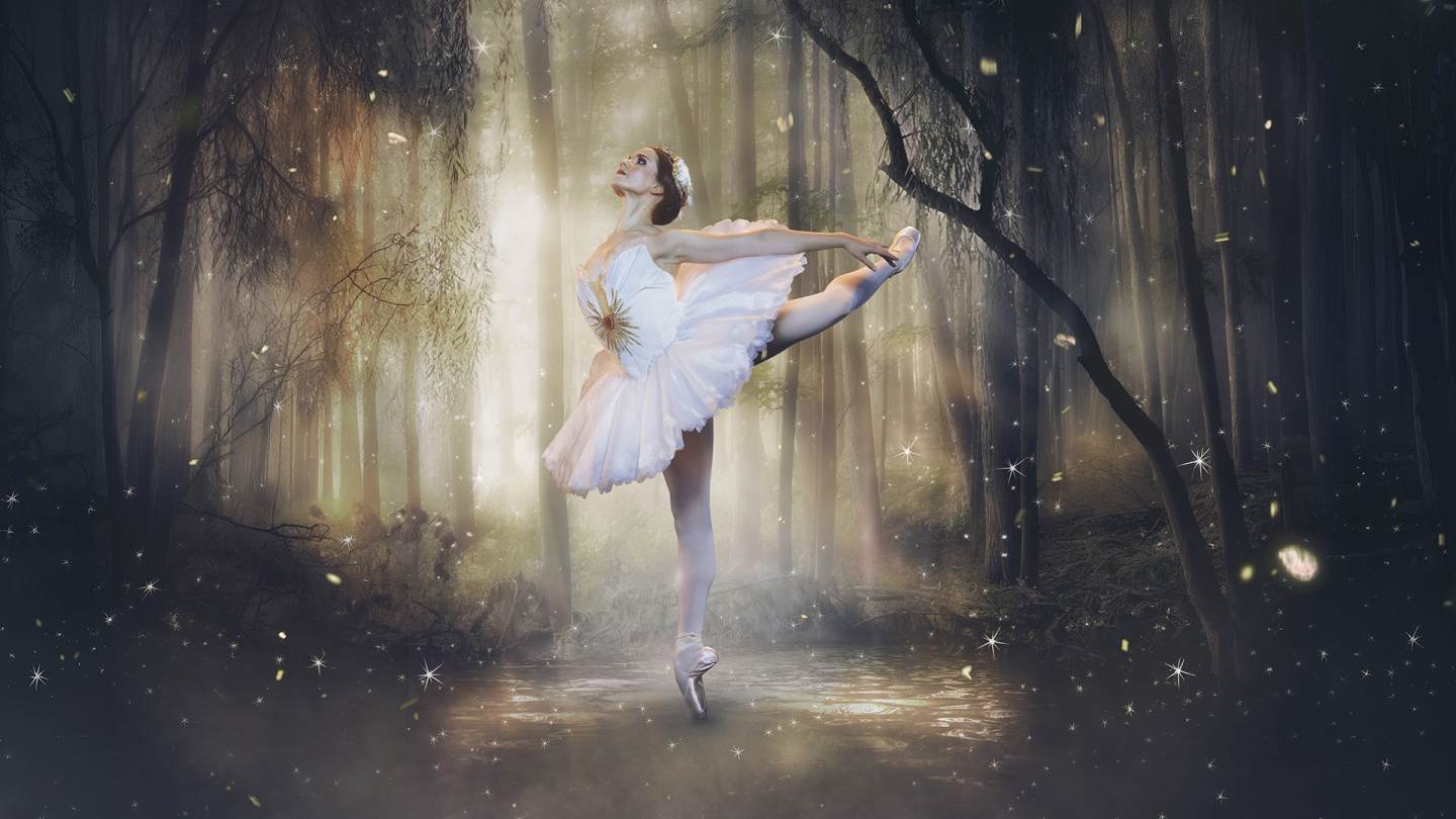 In the middle of a dark forest with soft sunlight, a dancer in a white feathery tutu poses in an attitude, with her back arched and her arms reaching to the back.