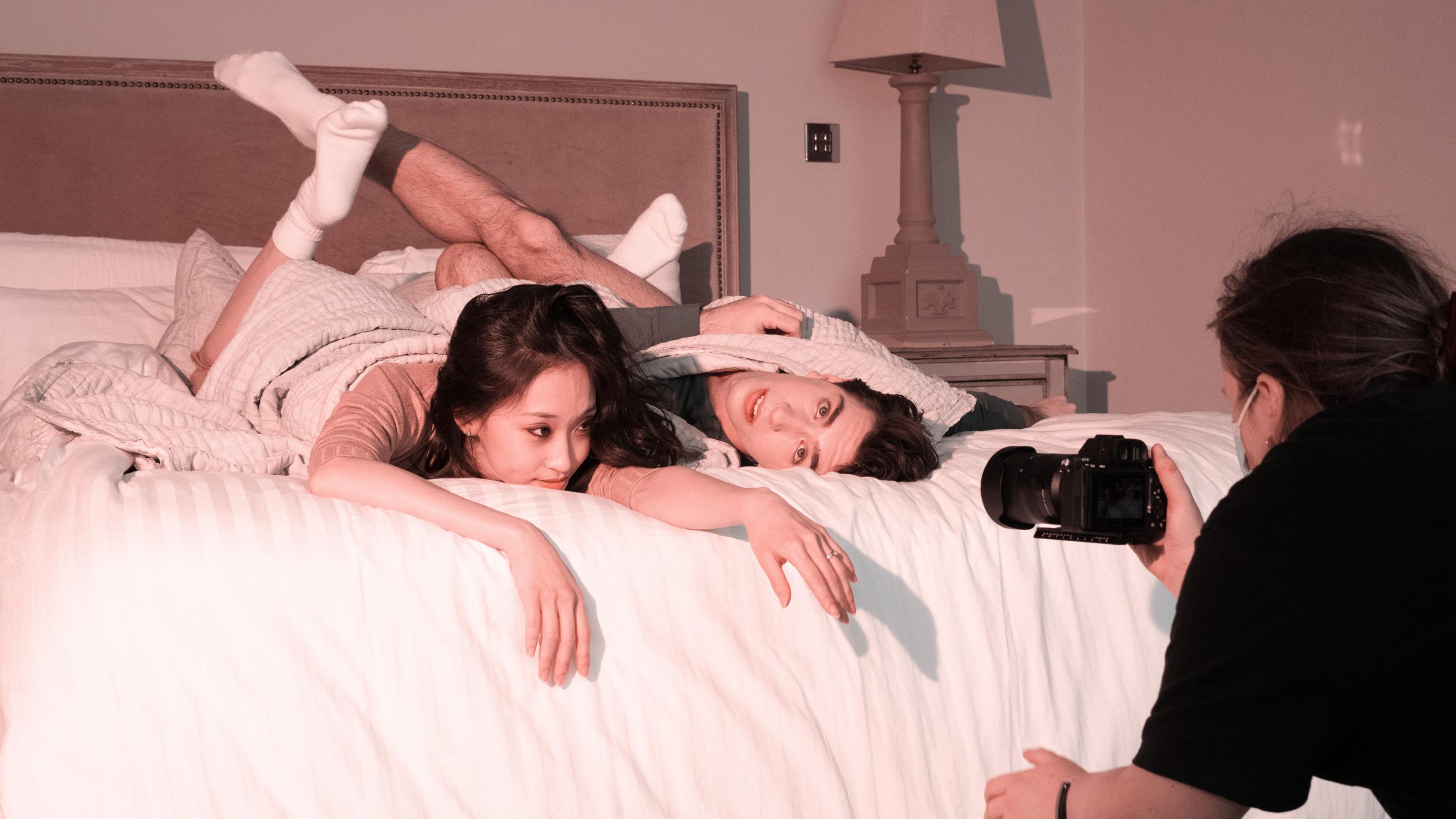 A woman and a man lay on a bed, the woman stares into a camera