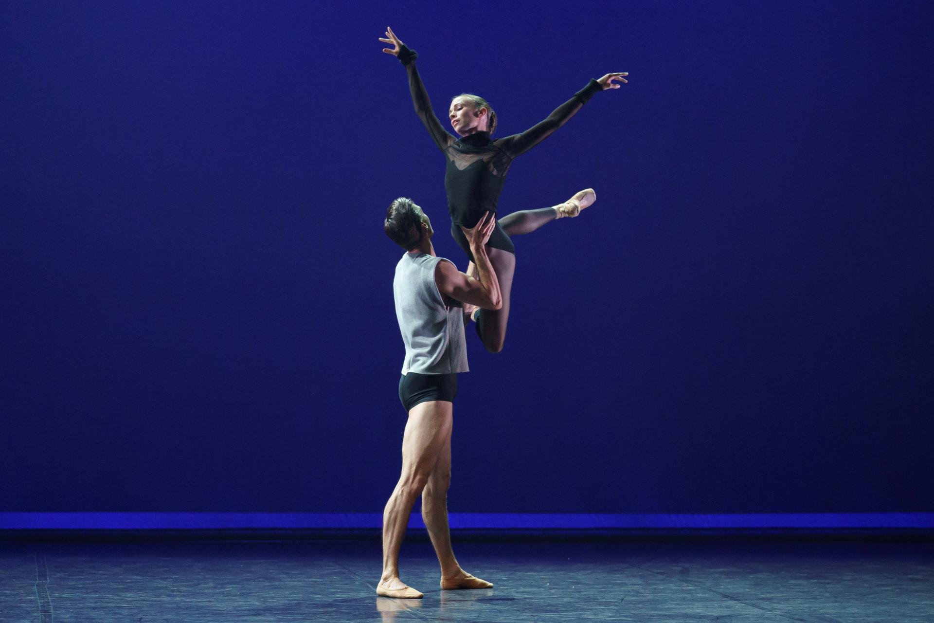 A female dancer in black leotard is lifted in the air by a male dance in black trunks and grey vest.