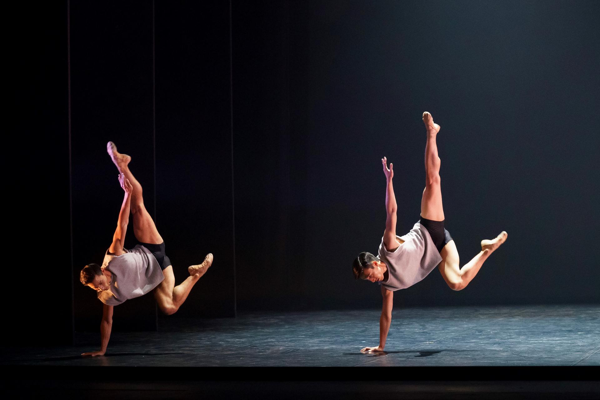 Two male dancers stand on one arm with opposite arm and foot pointed to the ceiling.
