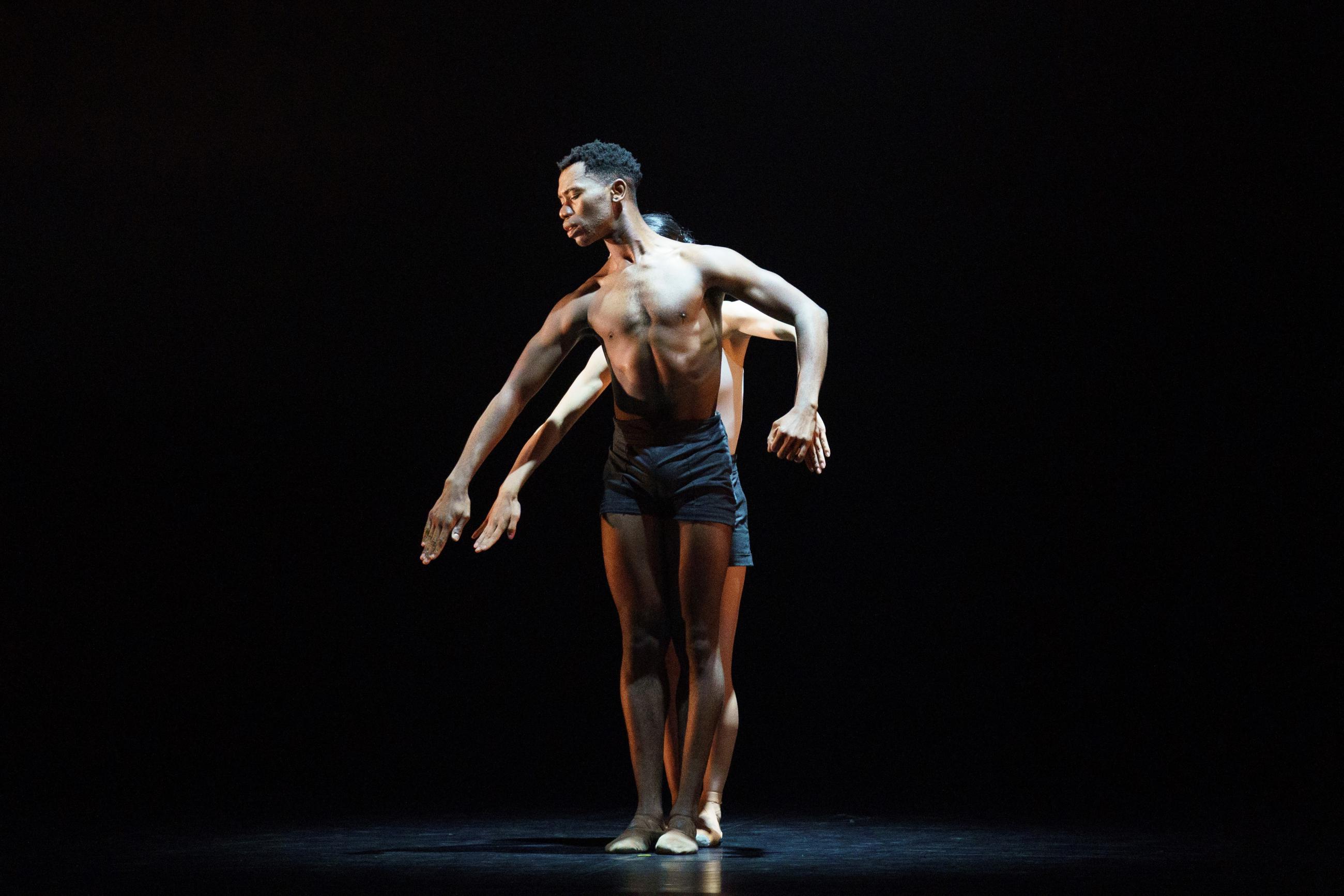 A male dancer stands topless as a female dancer mimics his movements from behind.