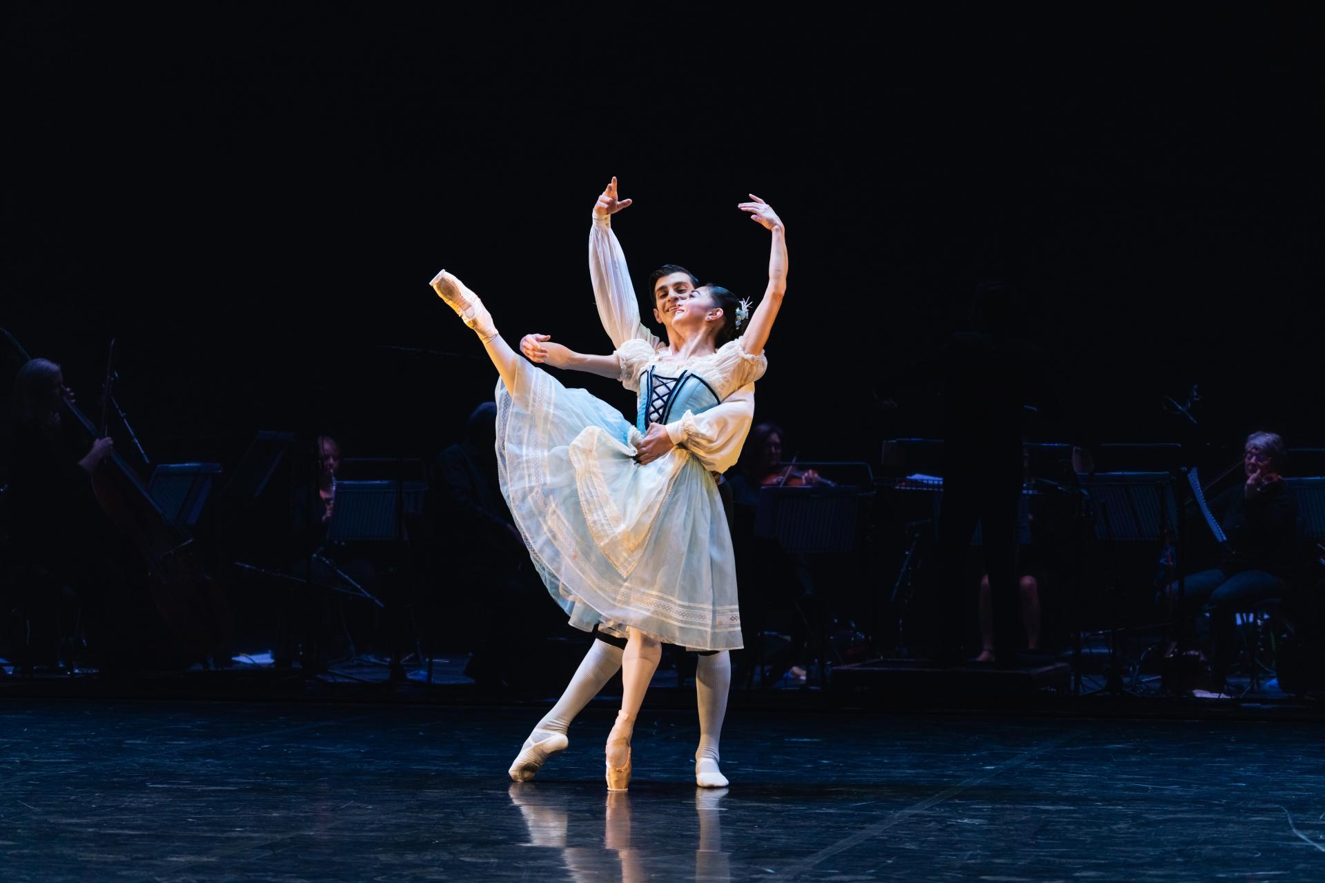 A female dance stand en pointe with her right foot pointed to the ceiling. A male dancer supports her from behind.
