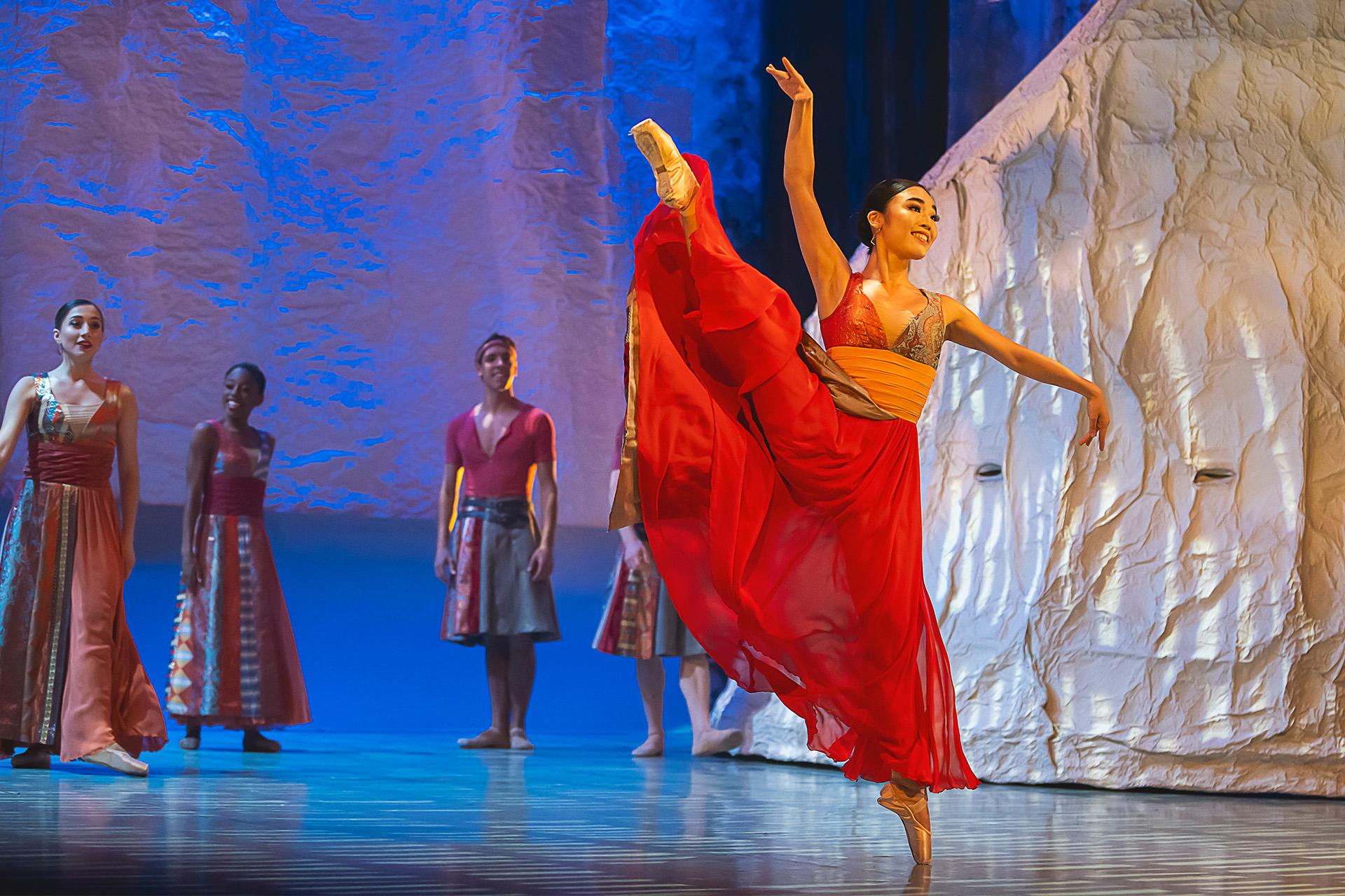 Female dancer in a flowing red dress raing her right leg above her head who en pointe on her left.