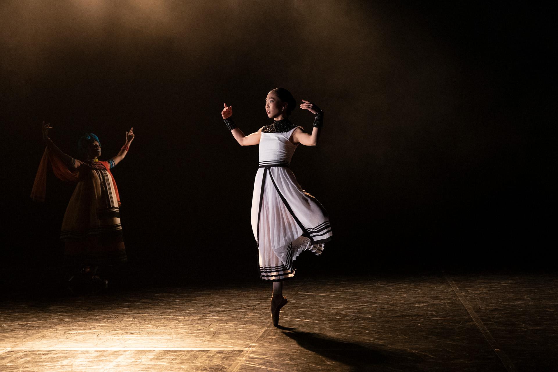 Dancer in a white dress with black details standing on a stage on one foot, en pointe, and arms lifted to shoulder height.