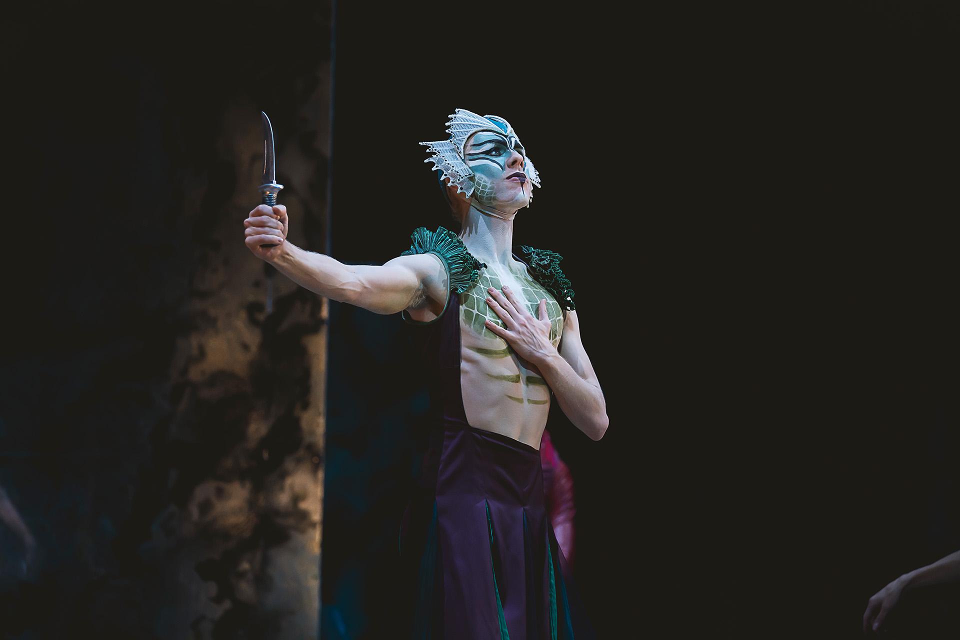 Lyr, Lord of the Sea, places one hand on his chest and holds the other out gripping a knife