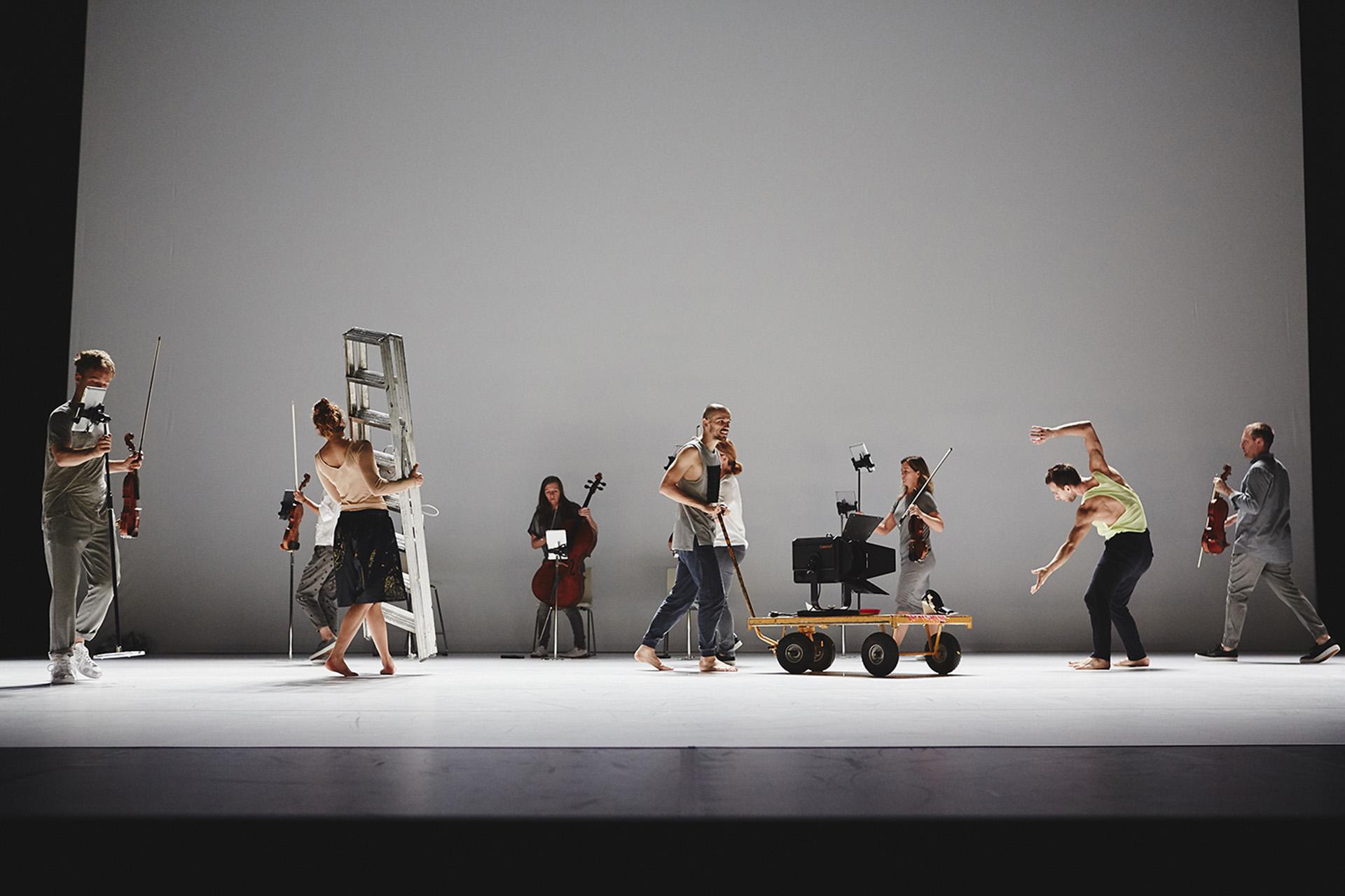 A group photo of a number of people on stage, one carrying a stepladder, another pushing a trolley, a third dancing, more people carrying and preparing stringed instruments