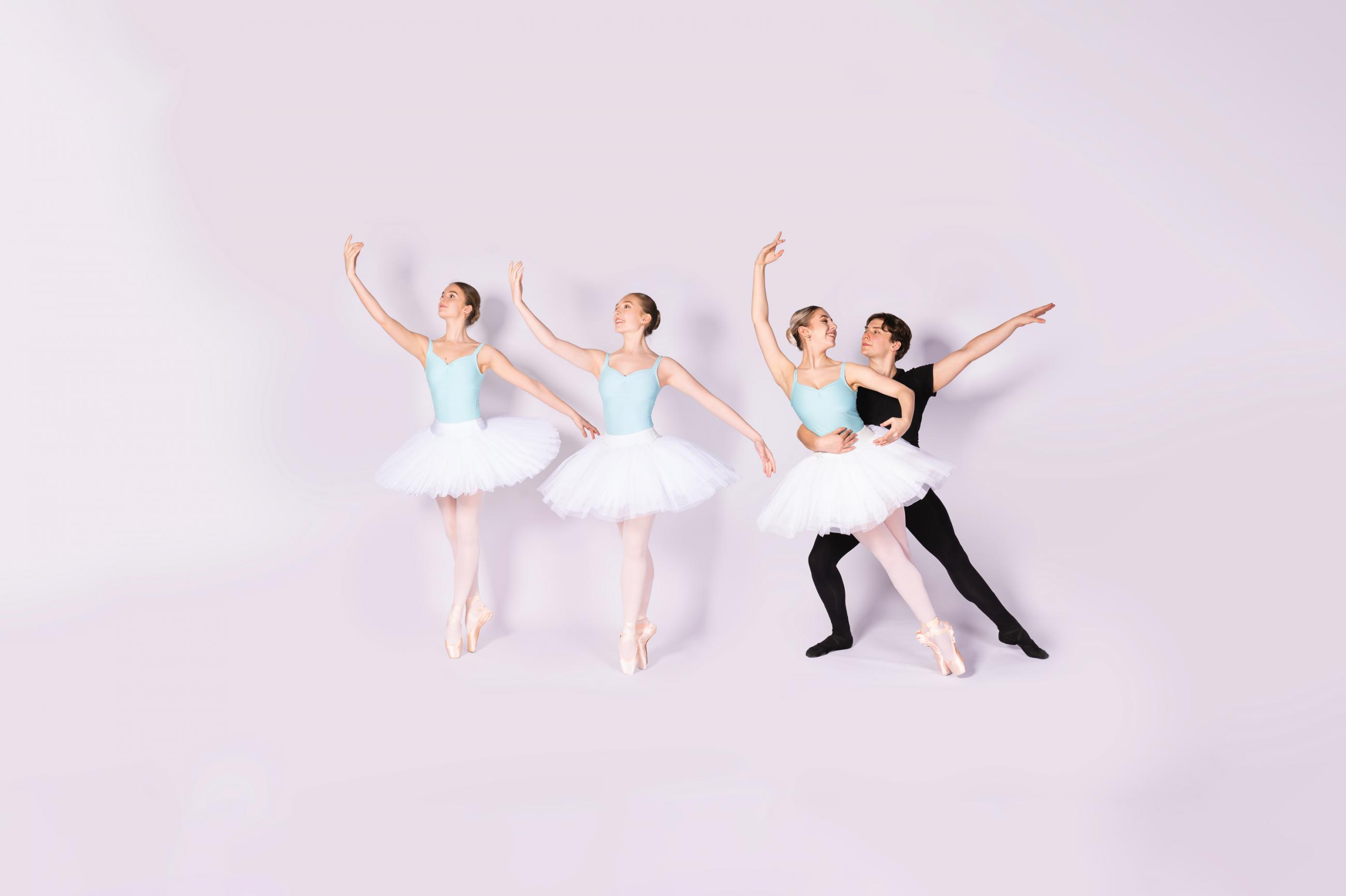 Three female dancers in white tutus stood on pointe. A male dancer holds one dancer sideways