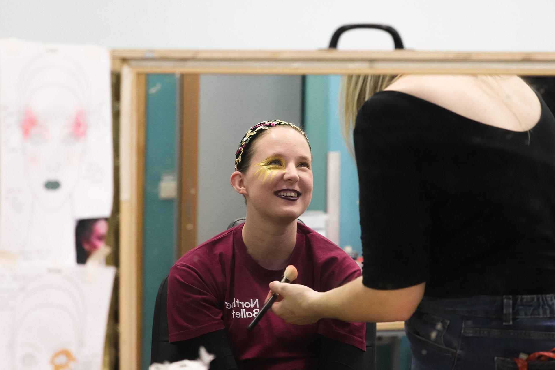 Photograph taken through a mirror showing a smiling performer have her make-up applied.