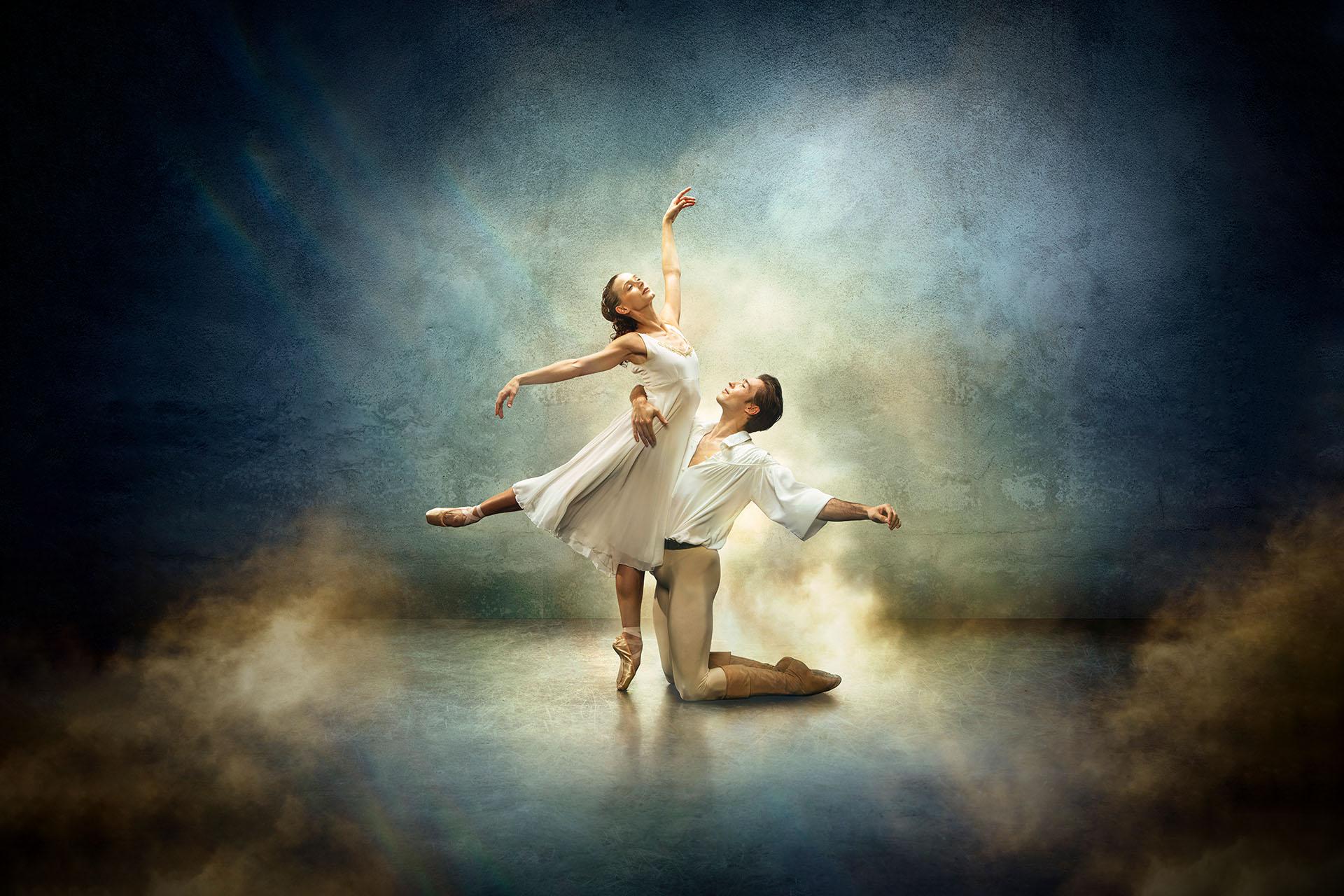 Star-crossed lovers Romeo & Juliet, her standing en pointe and him kneeling in front of her, both wearing white against a fantasy blue and yellow background.