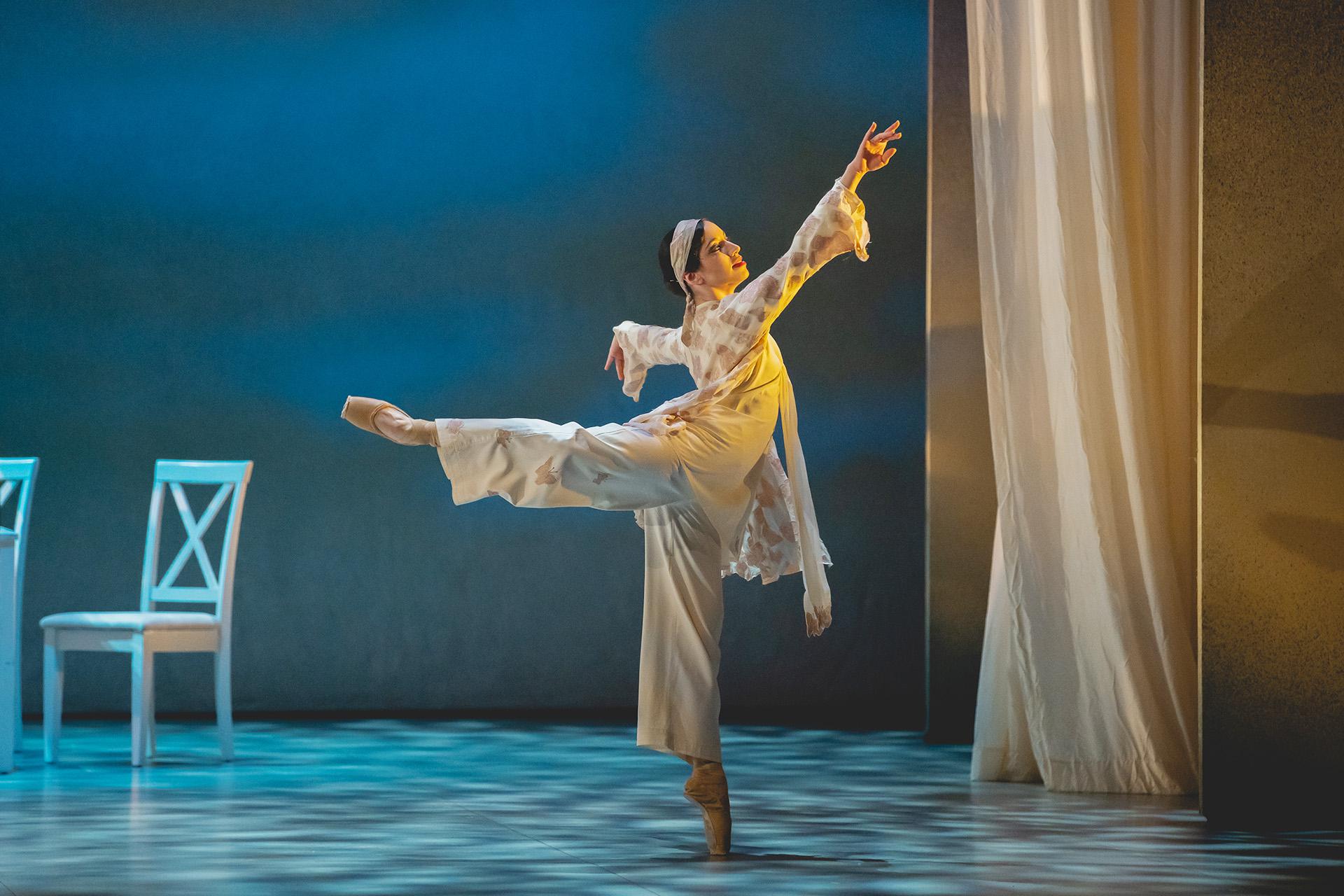 A production photo from The Great Gatsby of the character Jordan a suit of cream satin and wearing a headband, standing en pointe and arabesque