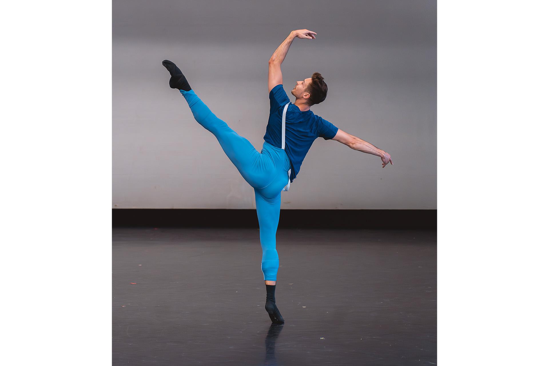Male dancer wearing blue stands on one leg, the other raised high, one arm above his head, and balancing with his other arm stretched out