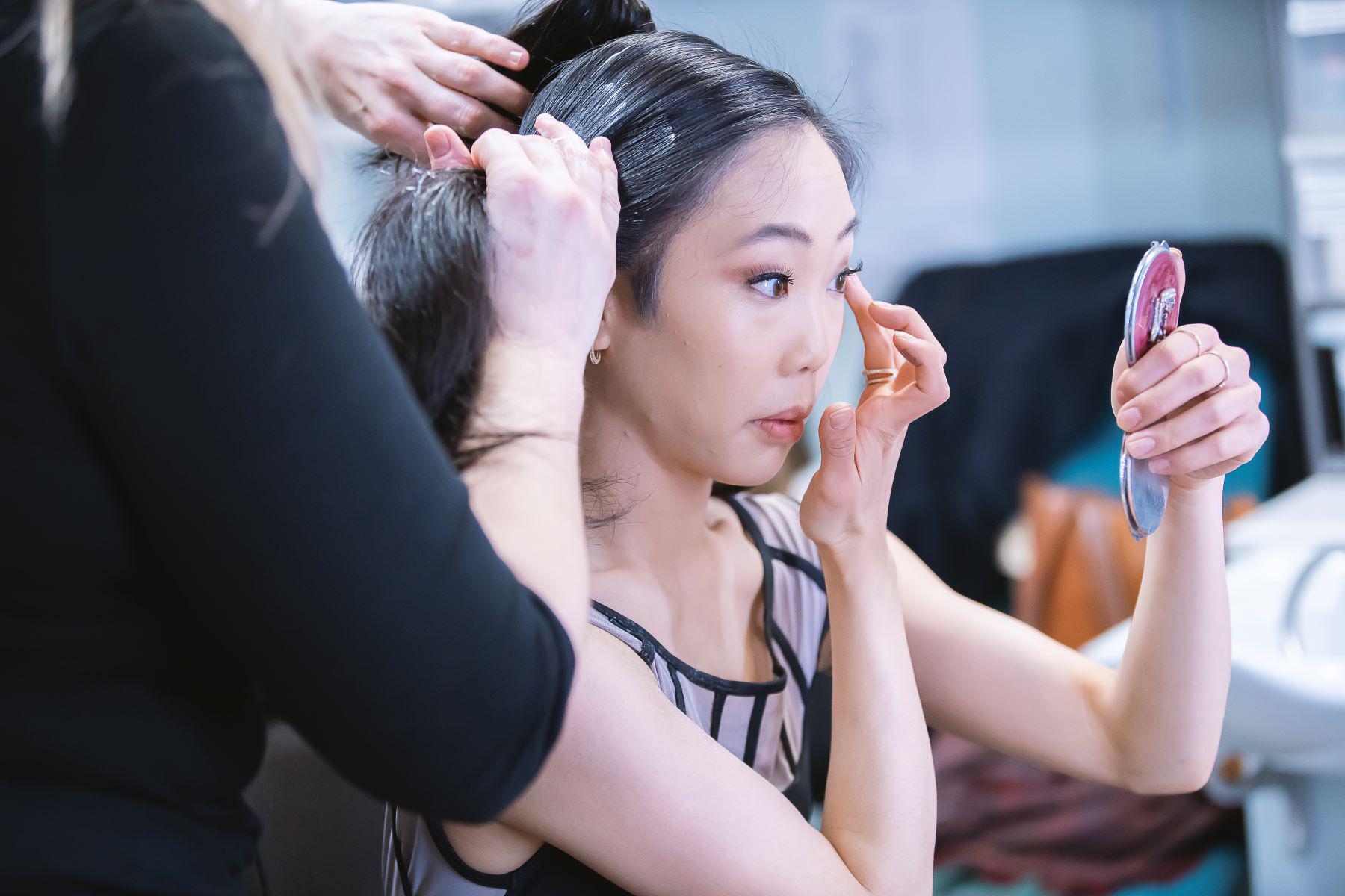 A female dancer applied makeup to her eyes whilst a hairdresser styles her hair