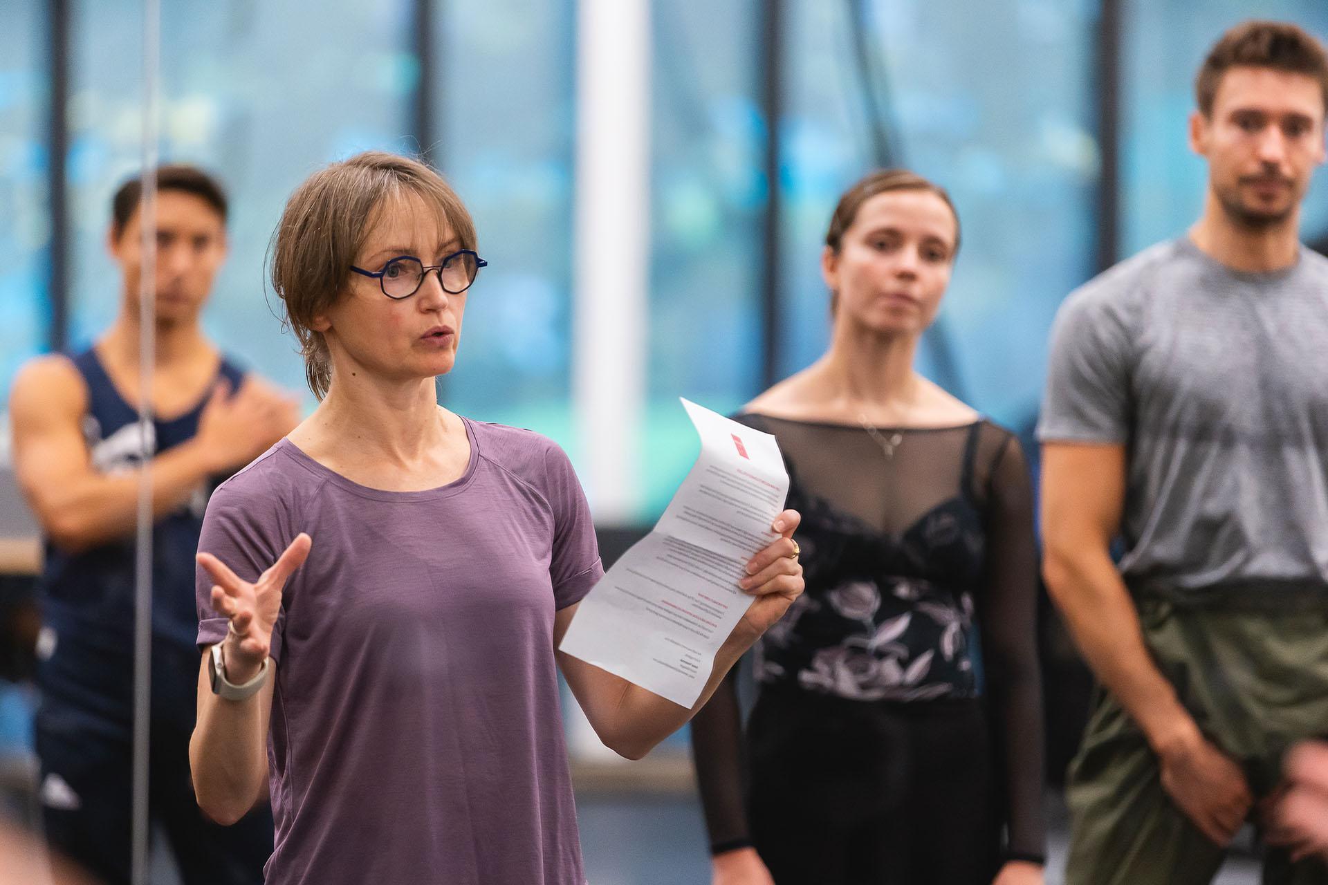 Repetiteur Larissa Lezhnina wearing round glasses and a mauve top and holding a letter while she addresses dancers in a rehearsal
