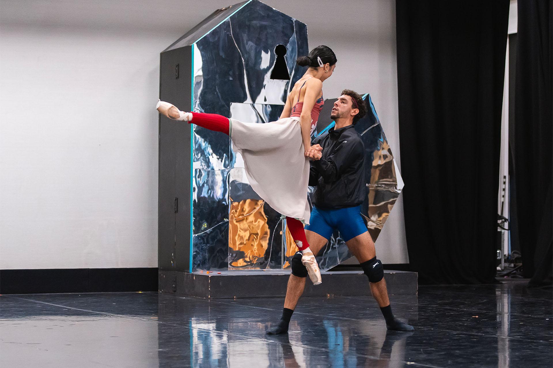 during rehearsals a male dancer stands supporting a female dancer whose legs are in arabesque but 40 centimetres above the ground, she's holding herself aloft by leaning on his arms which are bent to hold her weight