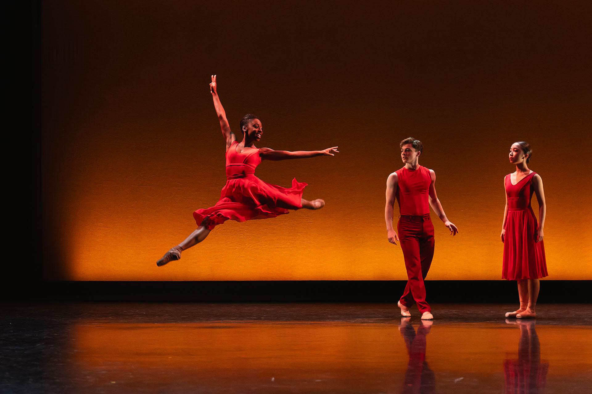 Generations, 2023, production, Aerys Merrill, Harris Beattie and Kaho Masumoto in Intimate Pages. Photos Emily Nuttall Female dancer wearing a red dress leap high with an arm stretched above her while two dancers dressed in red look on