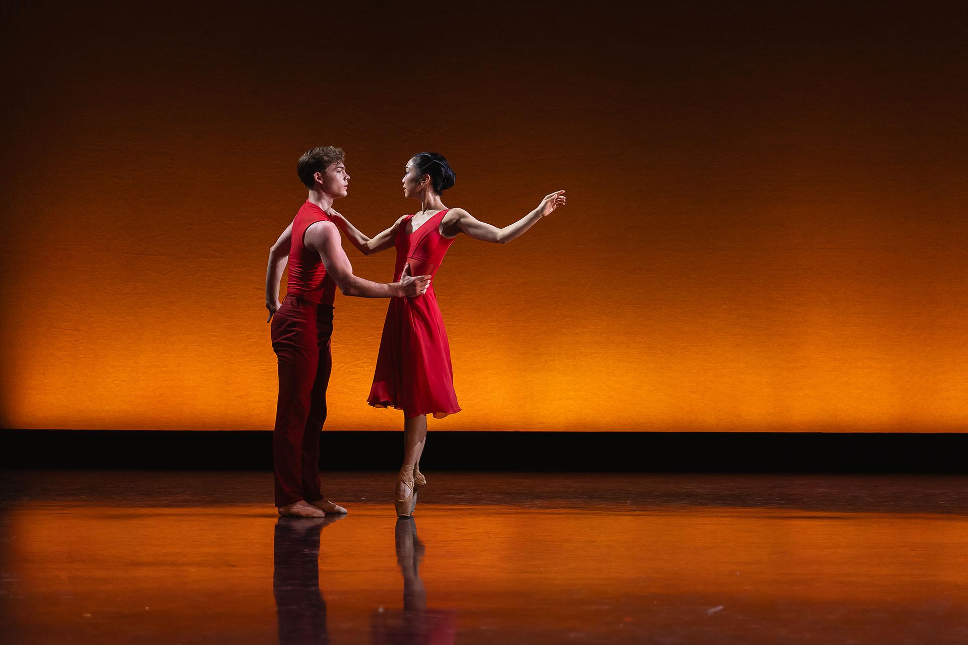 Two dancers wearing crimson, one holds the others waist and that person is en pointe, one hand on the other's shoulder