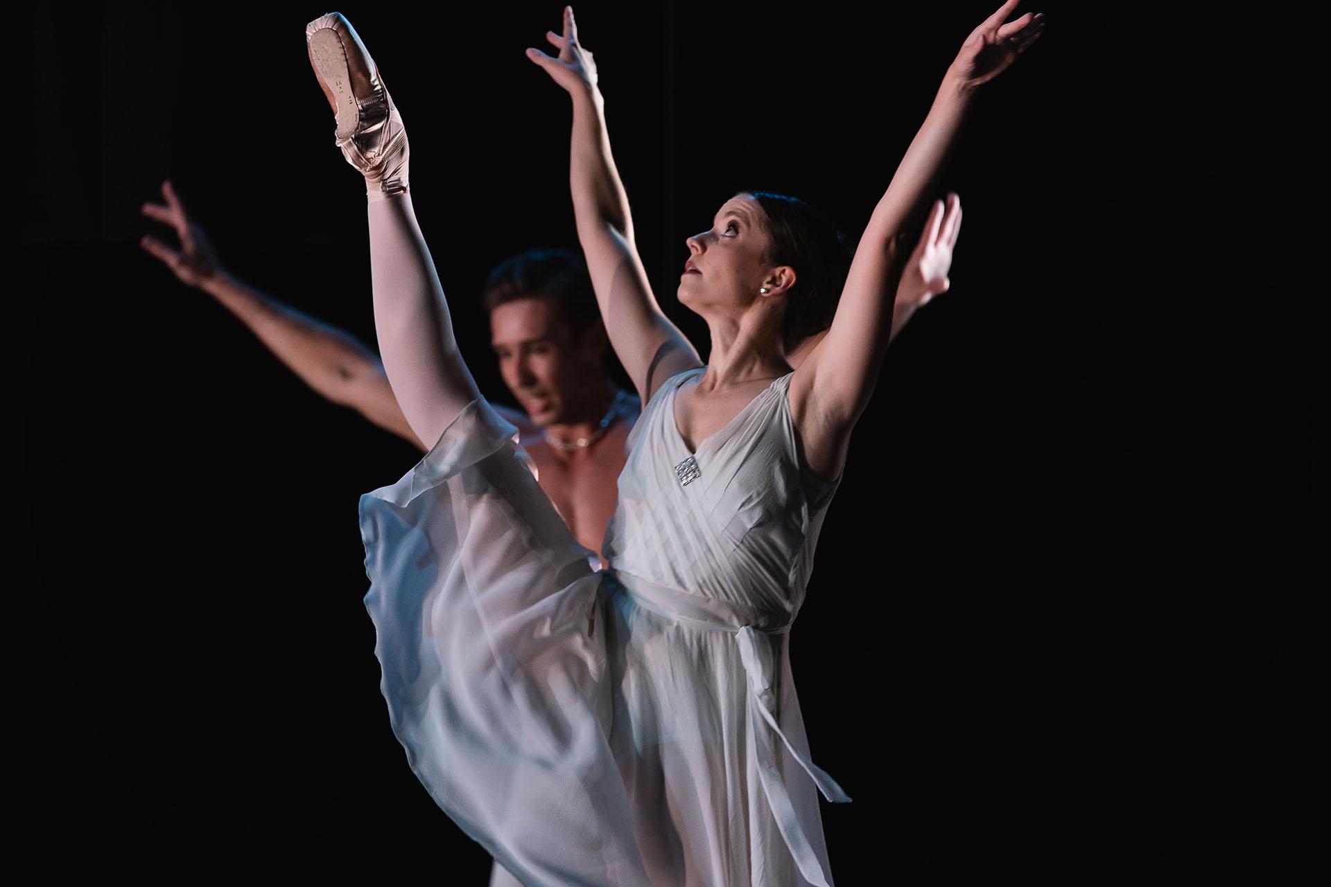 Female dancer against a black backdrop holds her arms above her head and one leg is raise near vertical , foot above her head