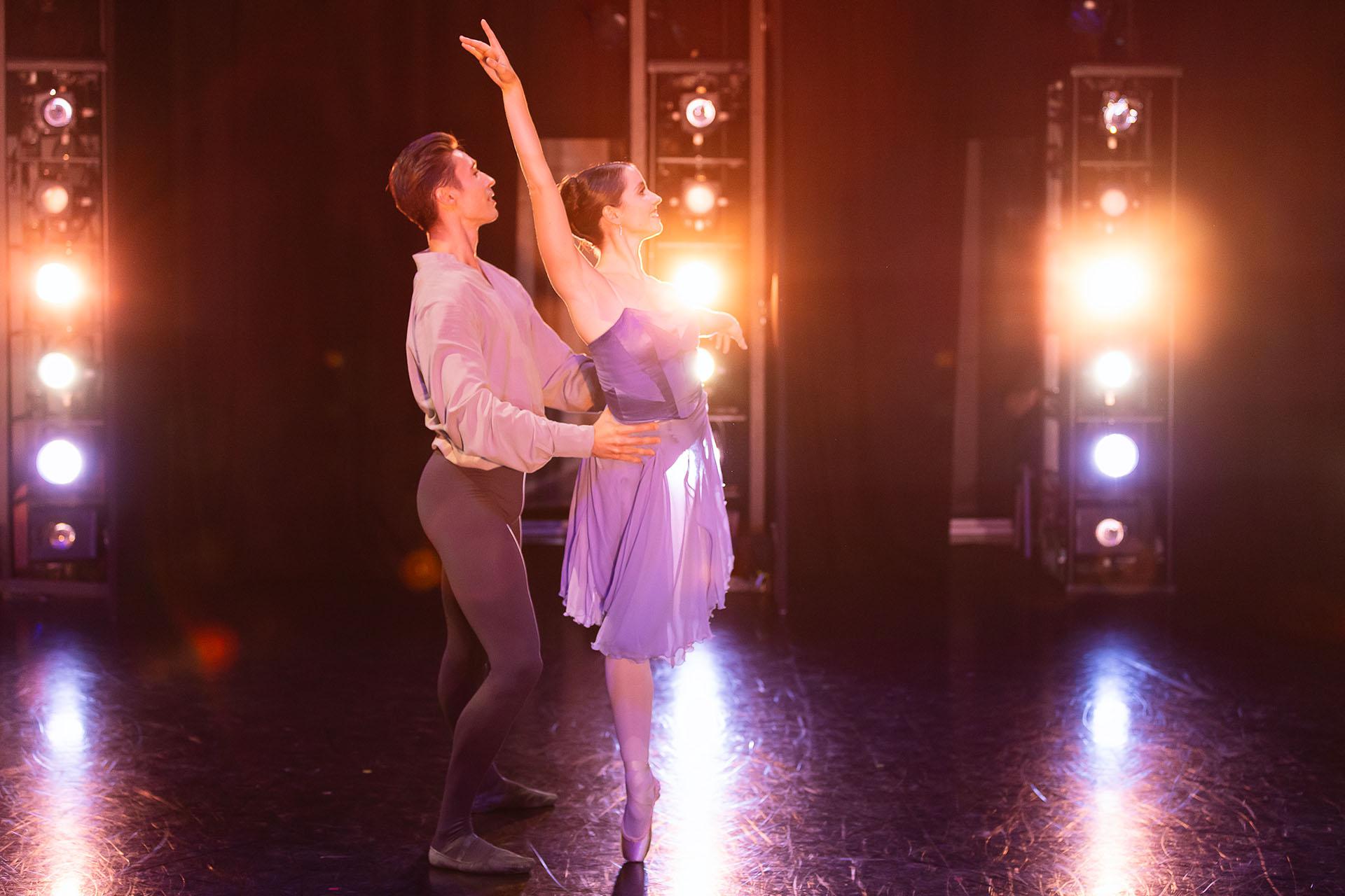 Looking across the stage the lists shine directly at us and in between we see a female dancer in a lilac dress standing on one foot with an arm above her head while a male dancer in grey tights and shirt holds her by her waist