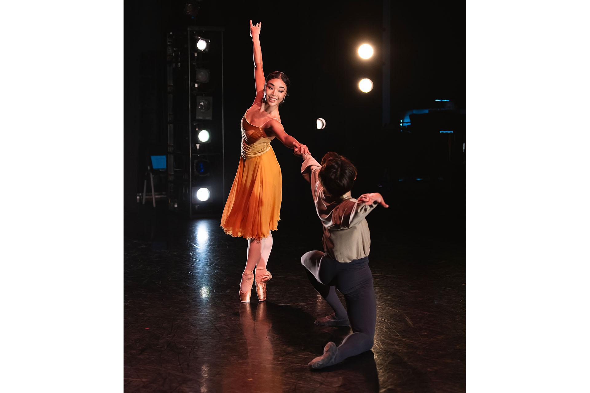 Generations, 2023, production, Kirica Takahashi and Jun Ishii in Joie de Vivre. Photos Emily Nuttall Female dancer wearing a vibrant orange dress stands on her toes and reaches across to a male dancer in grey tights and shirt who is kneeling on the stage and holding her extended hand