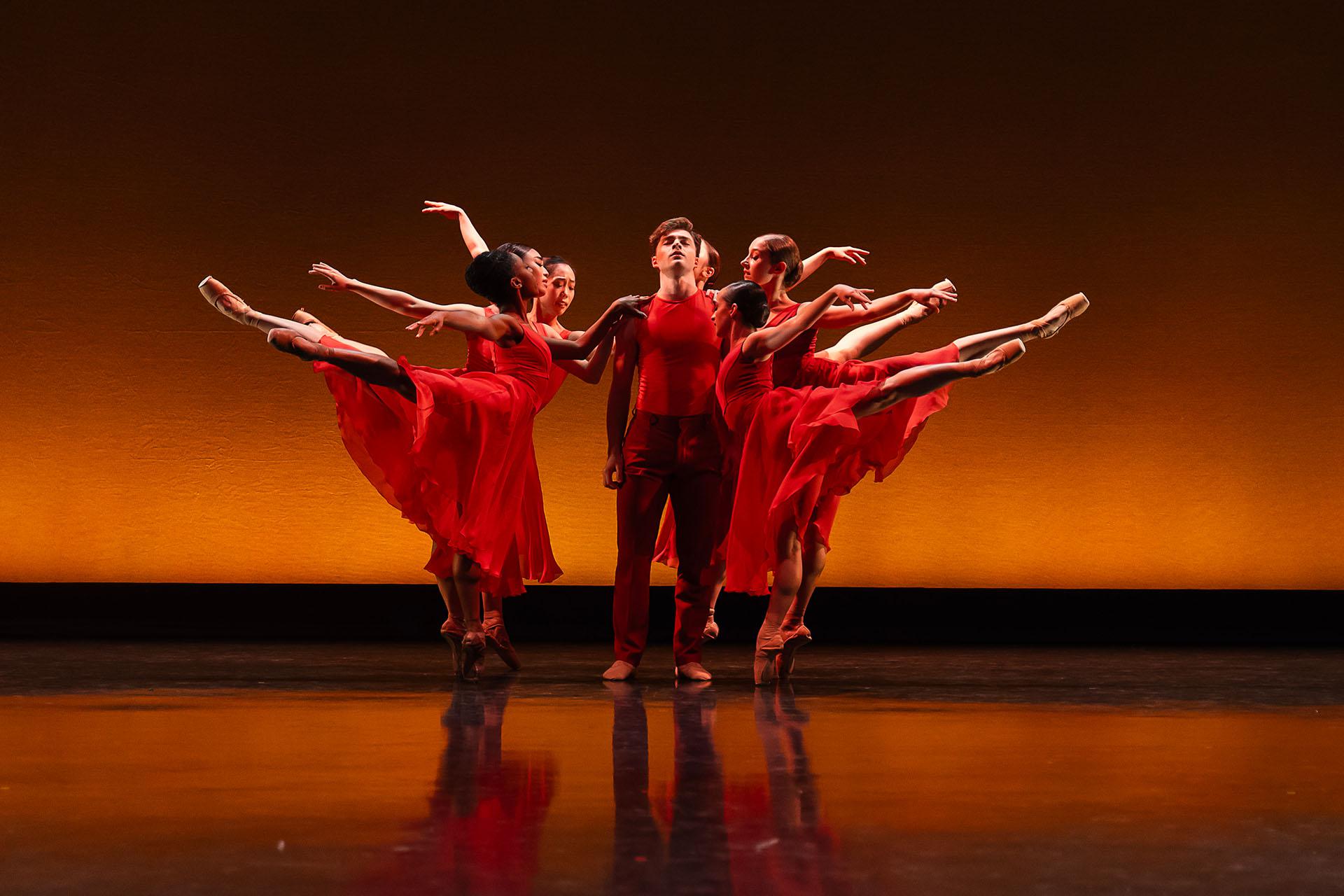 A male dancer in red is standing with arms by his side as female dancers also in red, stand arabesque around him