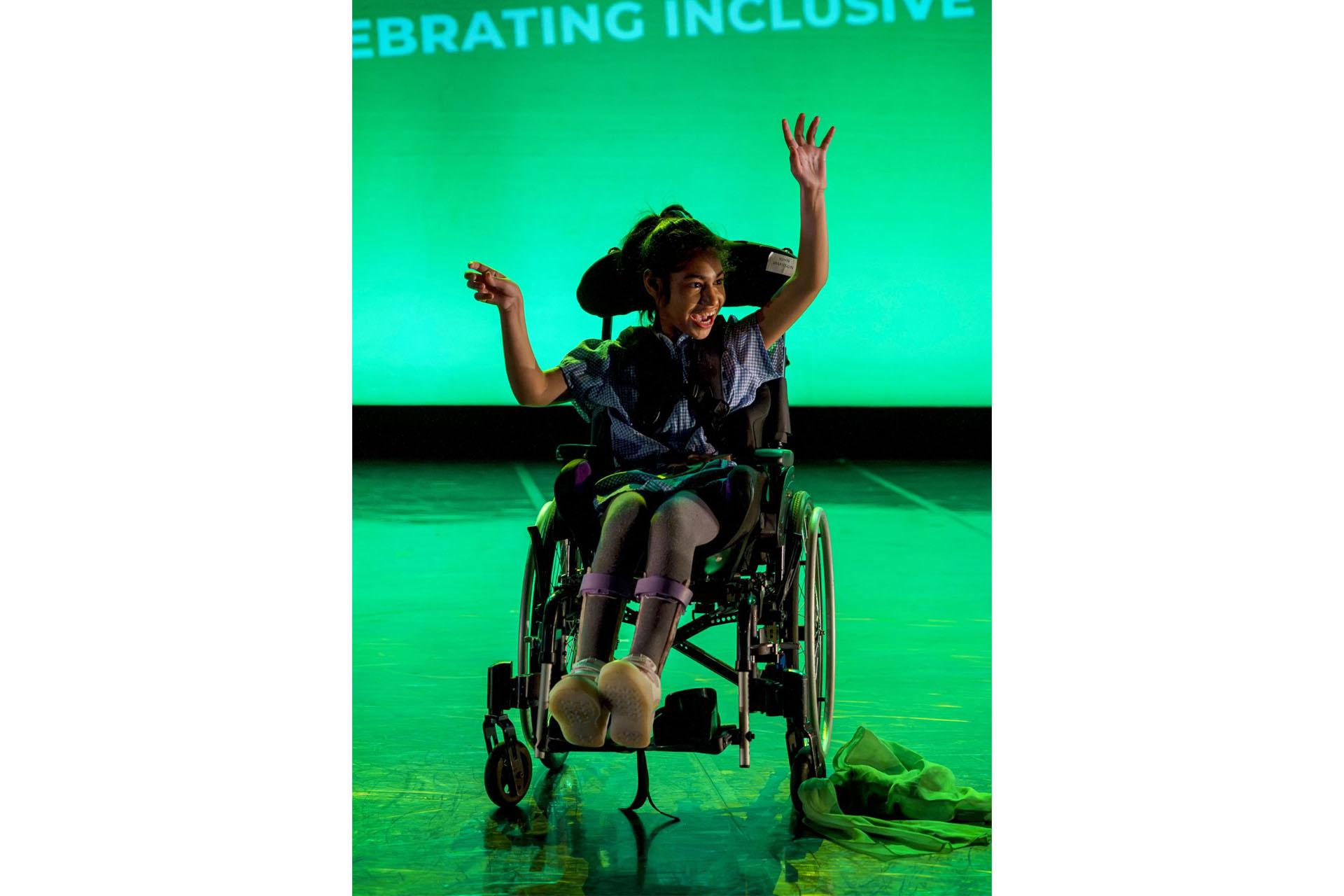 A young girl in a wheelchair throws her arms up in joy as she performs on a stage lit with green light