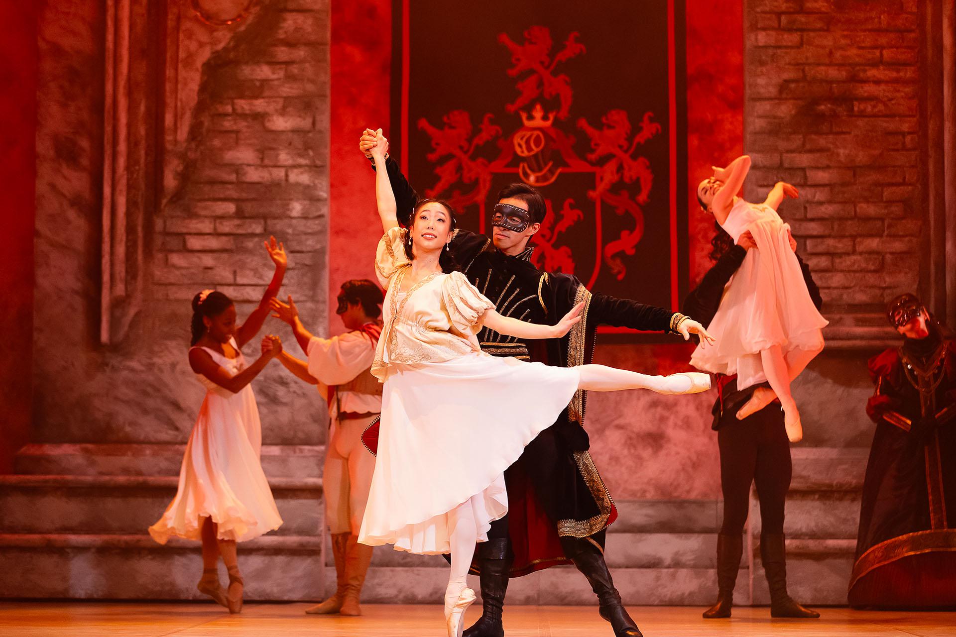 Paris dressed in a masque and in black provides Juliet with balance by holding her raised arm as she stands en pointe with a leg extended behind her