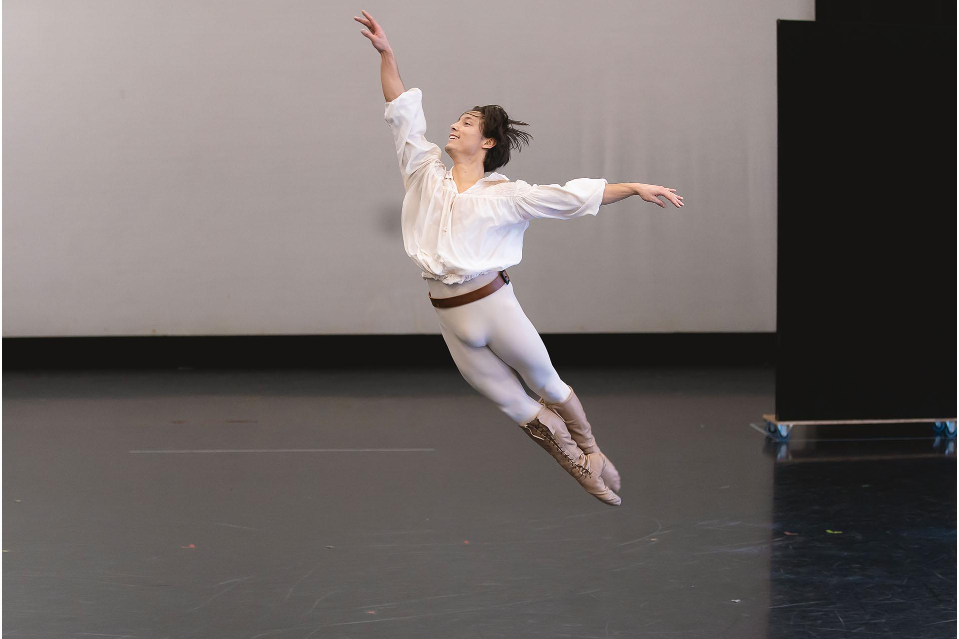 Man dressed in a white shirt and grey leggings leaps high with his feet together, one arm straight up above his head and the other straight behind him.