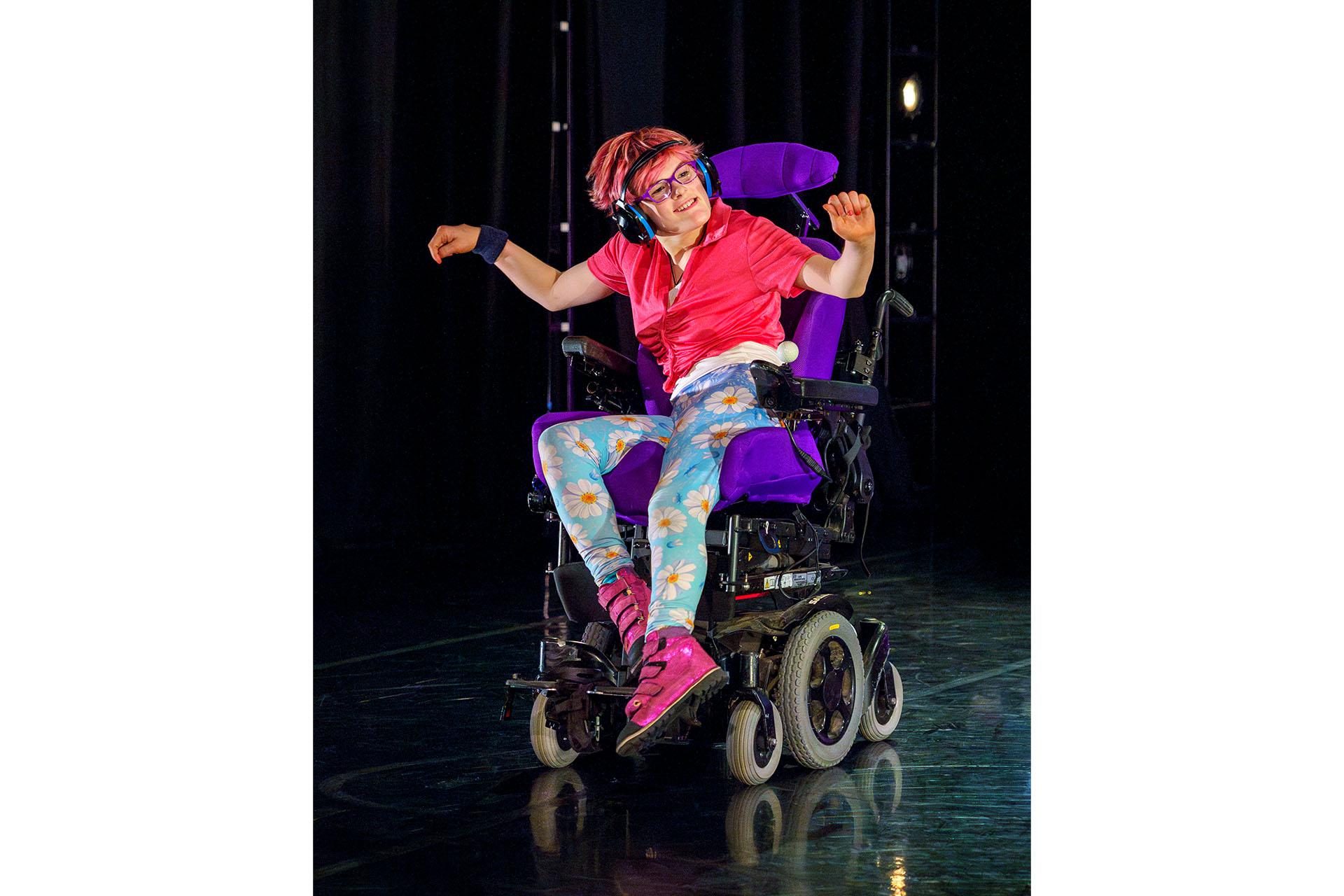 Woman wearing a pink to and flowery leggings riding a powered wheelchair, her arms are playfully out to her side and she has a broad smile on her face.