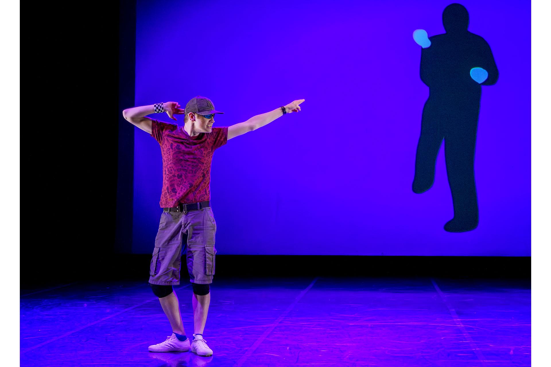 Against a blue background with a silhouette of a figure projected on it, a man in a baseball cap stands with his feet in first position, one hand to his head and the other stretched pointing to his left