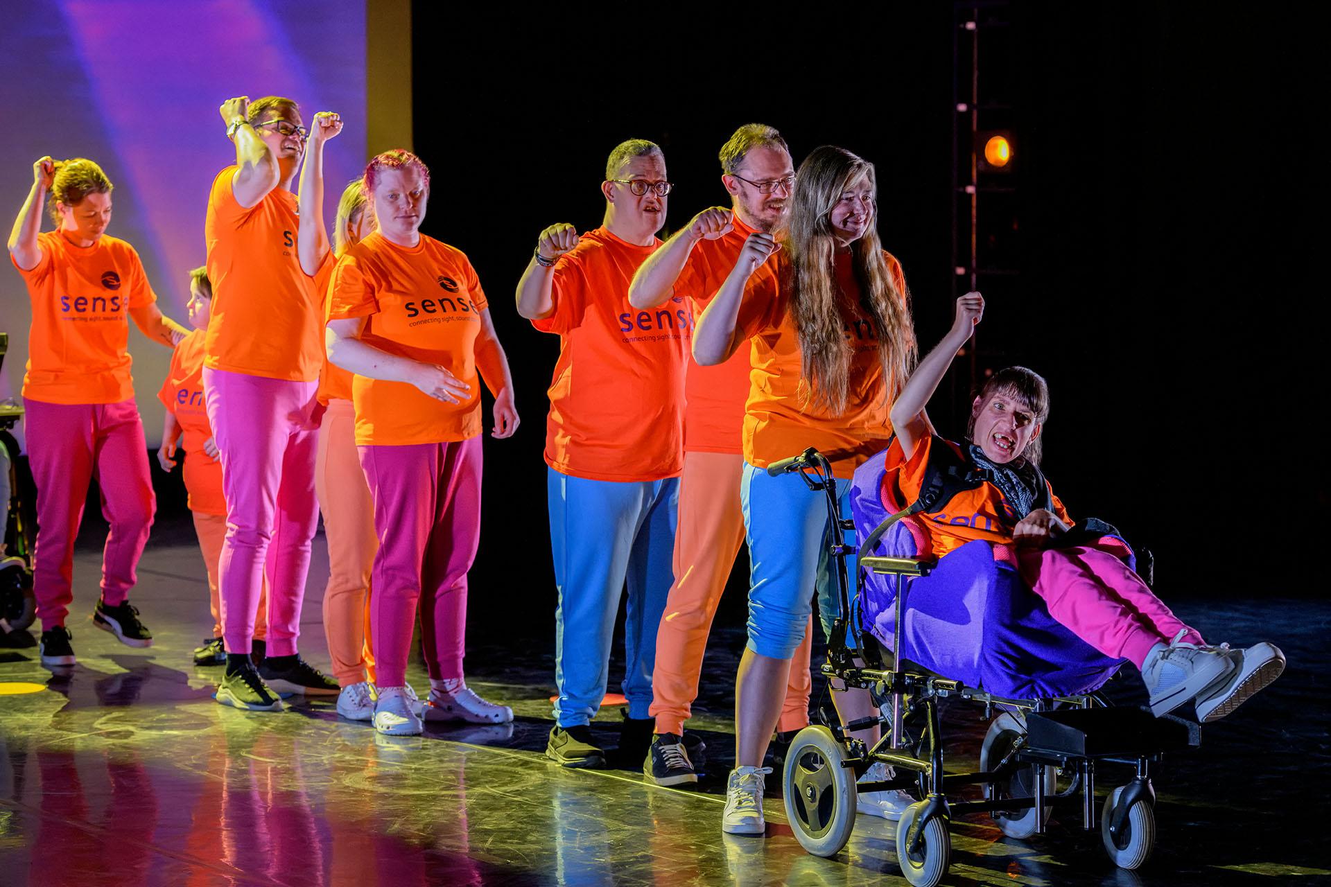 Group wearing orange T-shirts stand in a line, the right arm raised defiantly
