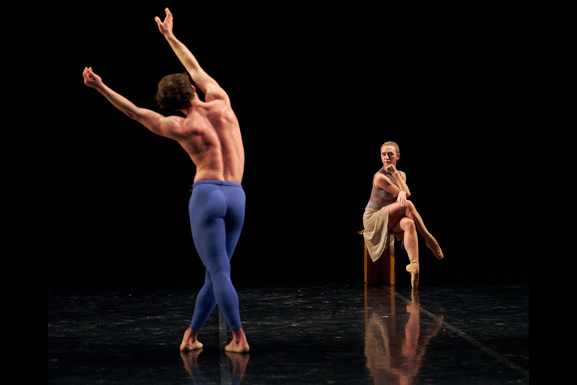 Dancer in the background sits on a stool resting watching another dancer walking towards her