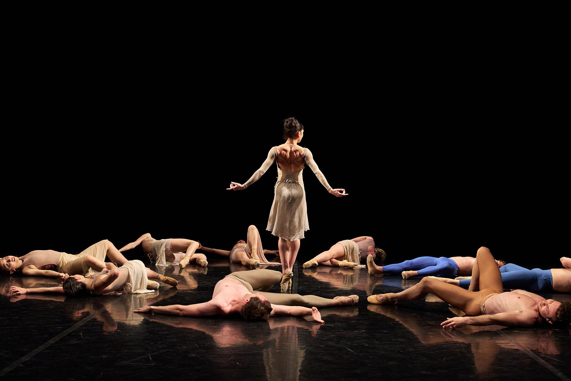 A group of dancers lie on the ground, apparently unconscious, while another stands and walks through the fallen bodies
