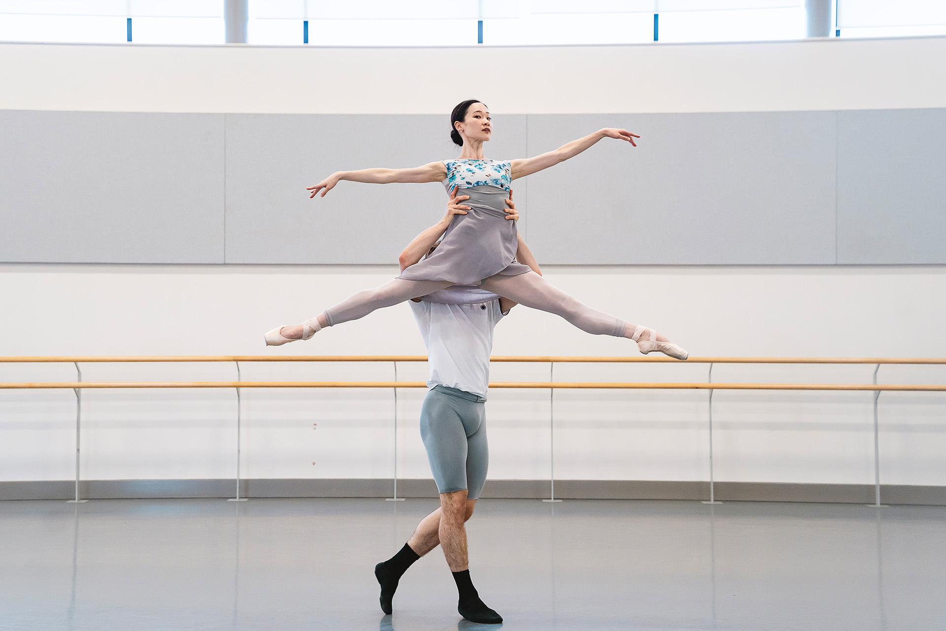 One dancer lifts another by the waist above his head, she holds her arms and legs stretched away from her body